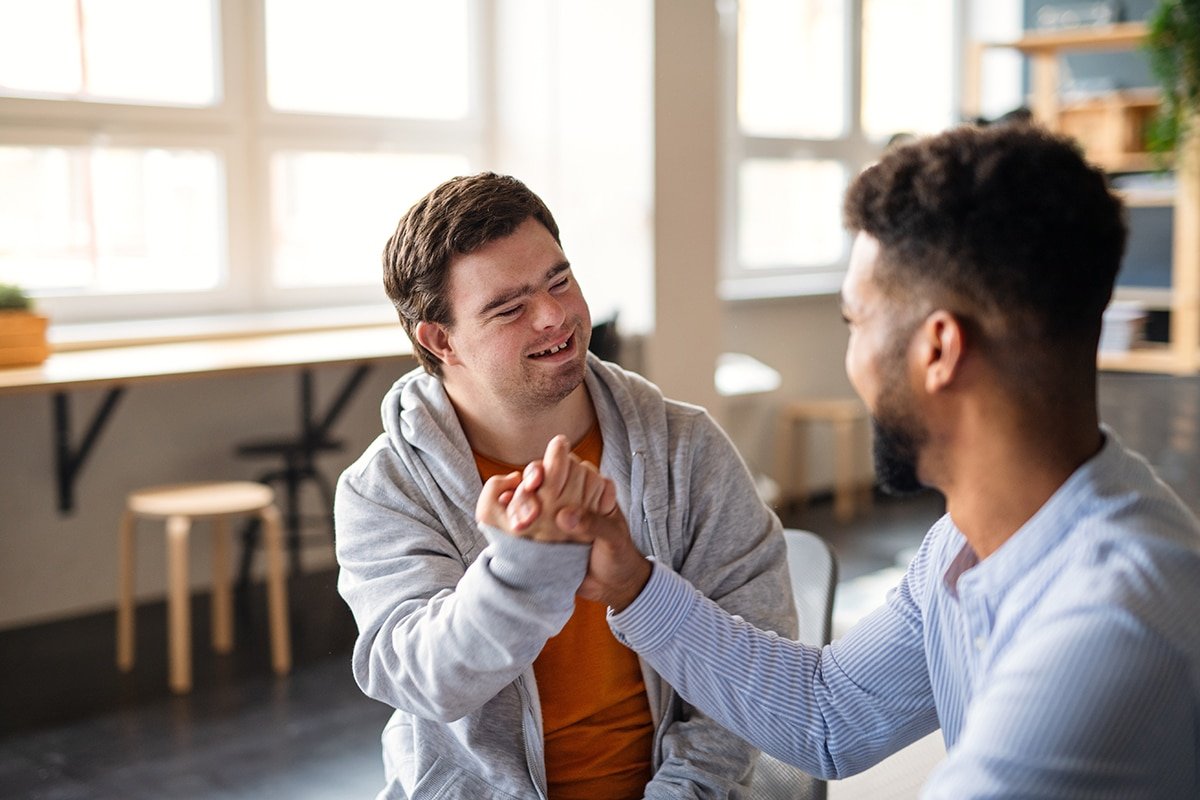 About Us 6 Stock image of Young happy man with Down syndrome with his mentoring friend celebrating success indoors at school